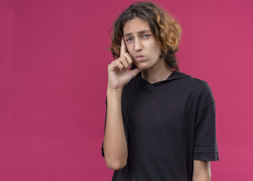 pensive guy with long hair in black t-shirt put hand on his cheek on pink background