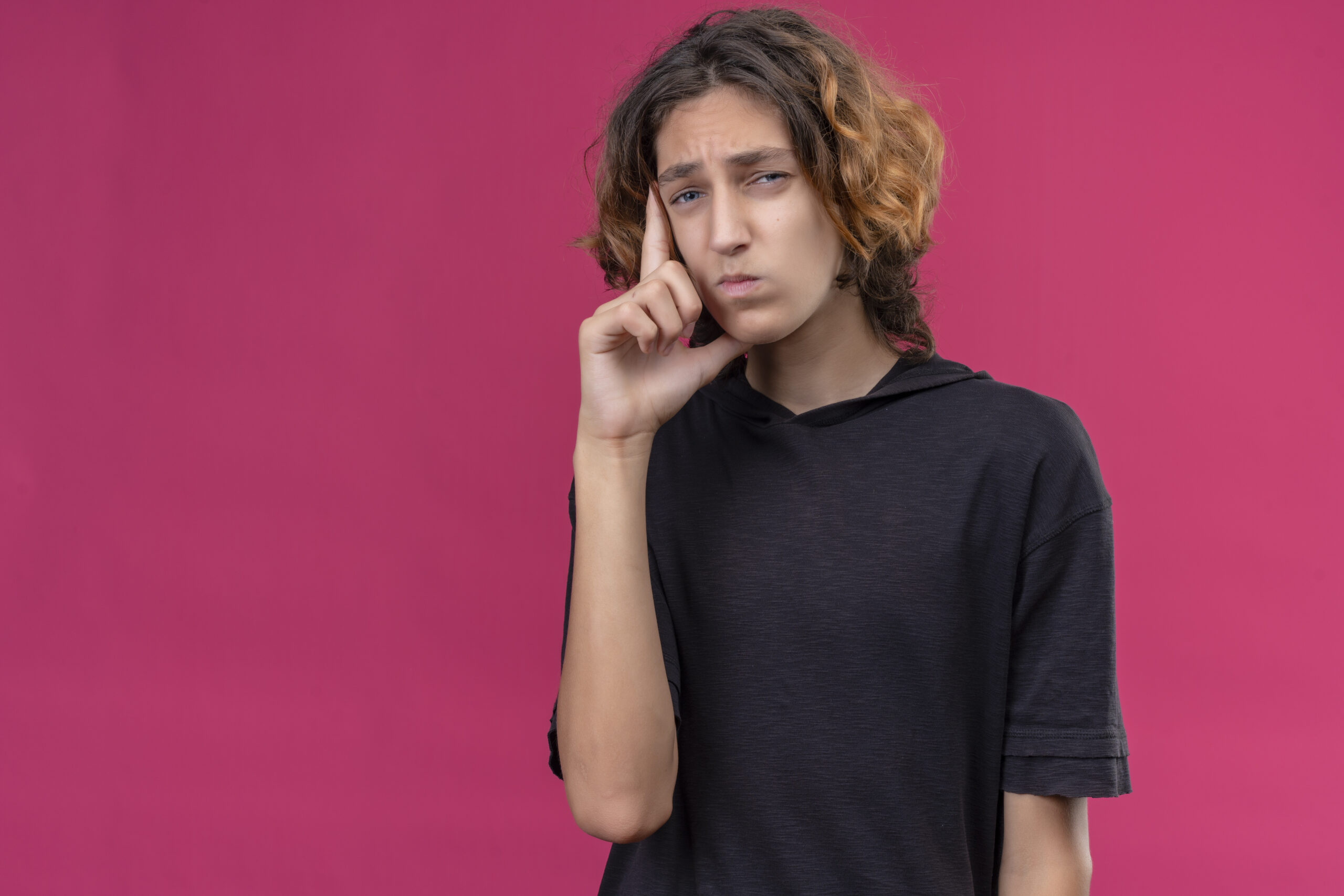 pensive guy with long hair in black t-shirt put hand on his cheek on pink background