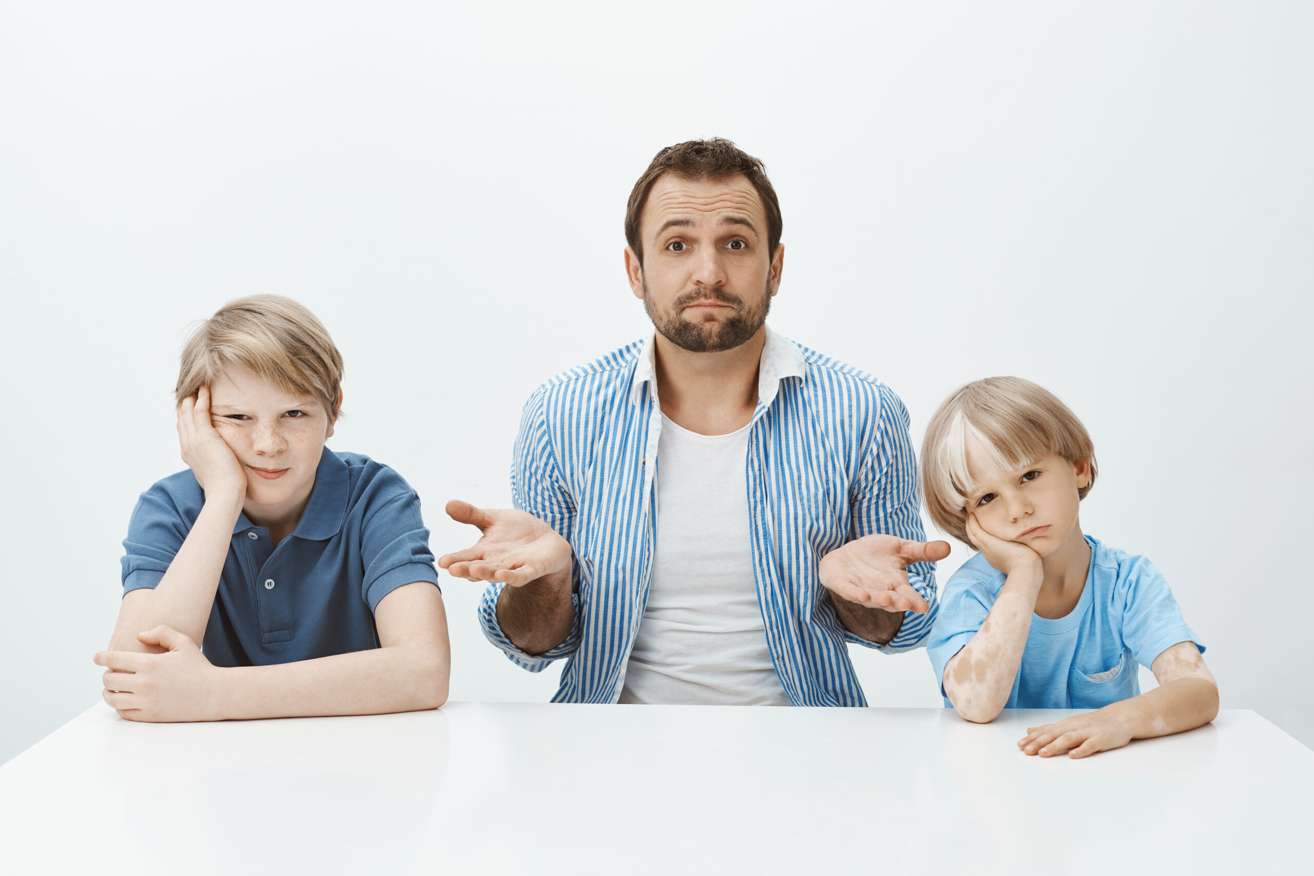 Portrait of questioned good-looking european father sitting at table with bored and upset sons, shrugging with raised palms, being clueless how to raise boys alone, sitting confused over grey wall