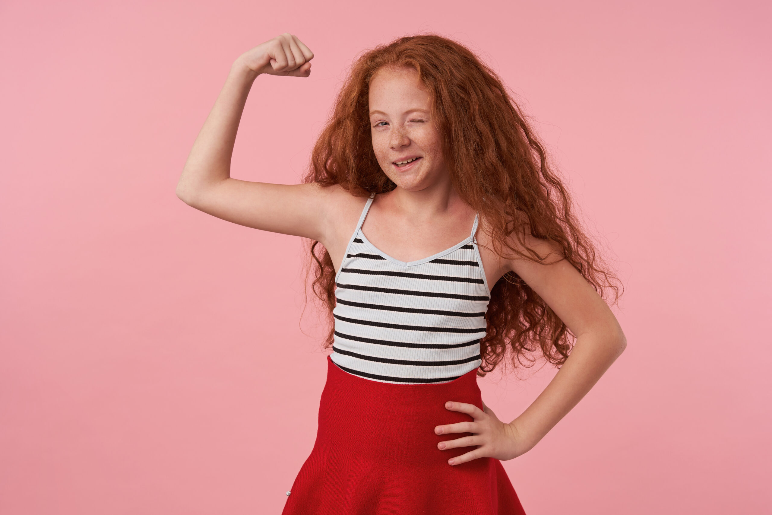 Posititve lovely female kid with long foxy hair posing over pink background in red skirt and striped top, winking cheerfully to camera and raising hand to show her power